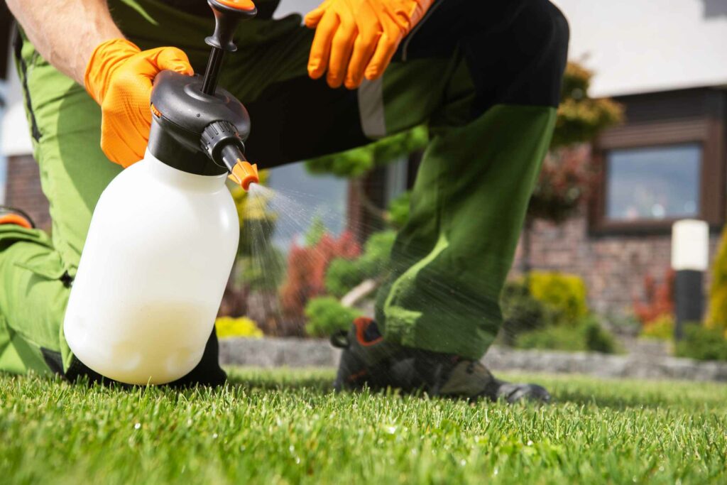 A man in green pants and orange safety gloves sprays down a lawn with a pesticide chemical